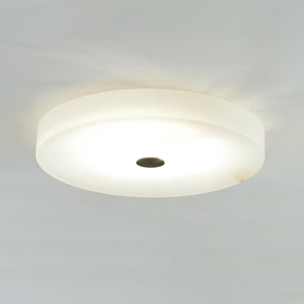 Wide view of the Olaean flush mount switched on, dark bronze cap centred in the glowing alabaster disc.