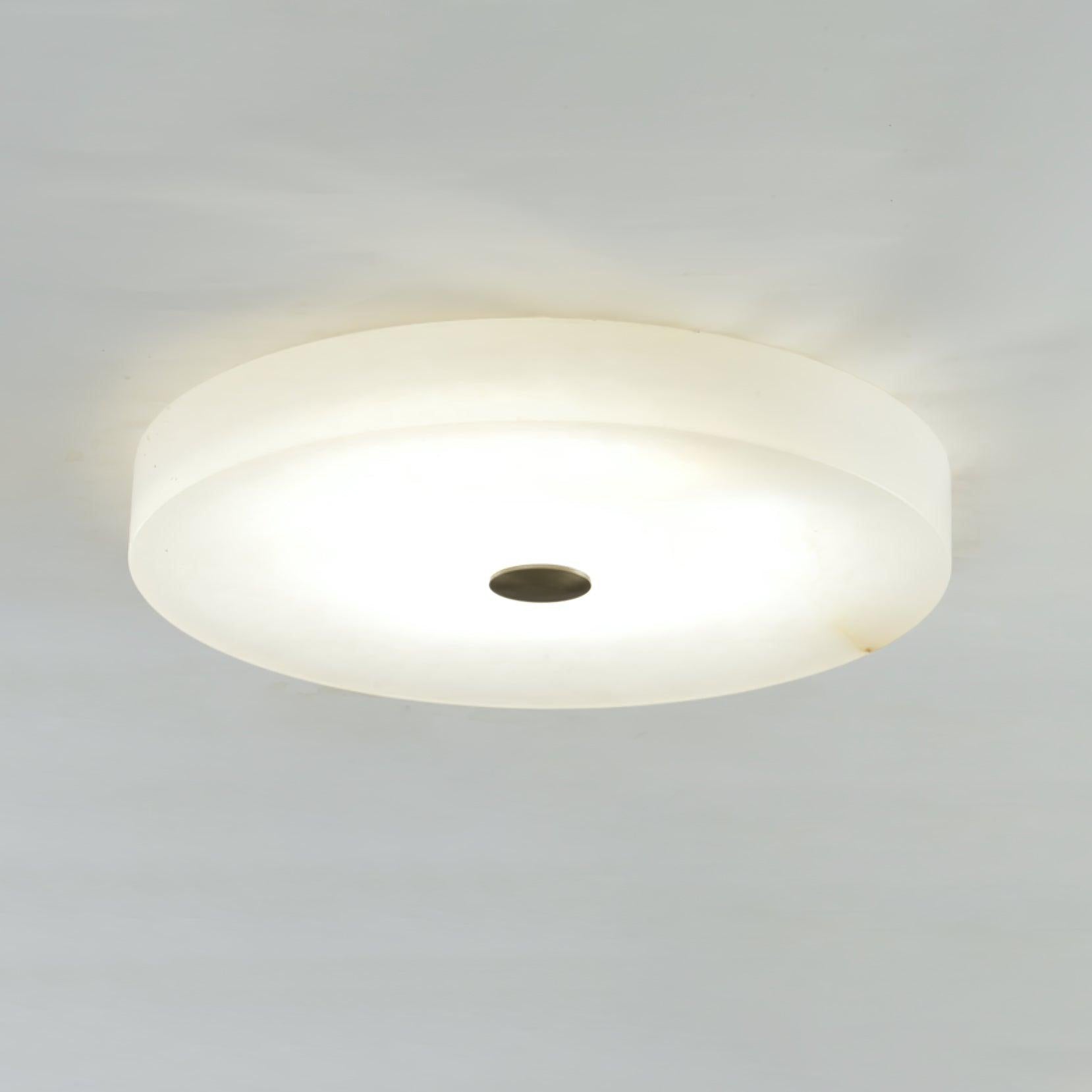 Wide view of the Olaean flush mount switched on, dark bronze cap centred in the glowing alabaster disc.