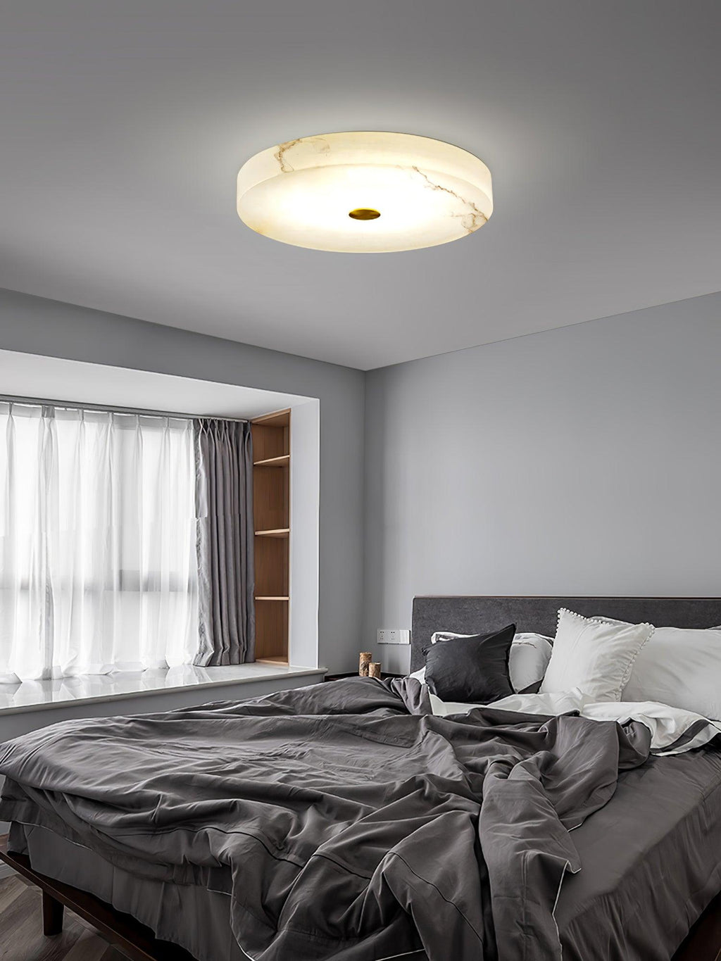 The lit Olaean flush mount on a grey bedroom ceiling above a bay window and rumpled bedding.