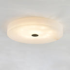 Angled view of the lit Olaean alabaster flush mount, showing the shallow drum profile and dark cap.