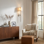 Beside a curtained window and cream armchair with ottoman, this Clérachic sconce in brushed brass lights a console table.