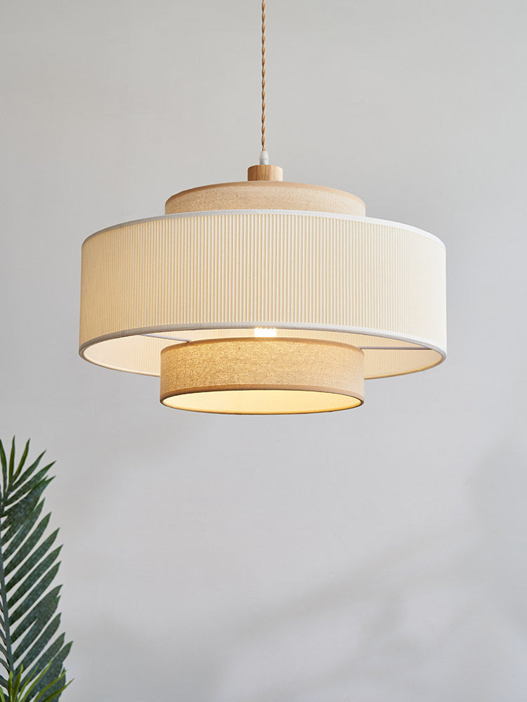 Against a plain grey backdrop, the white pleated band and beige drum of this Clérachic pendant catch the light, a palm leaf beside it.