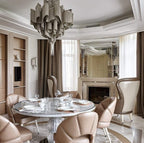 A round table and fireplace sit beneath this lit silver Clérachic chandelier in a beige dining room.