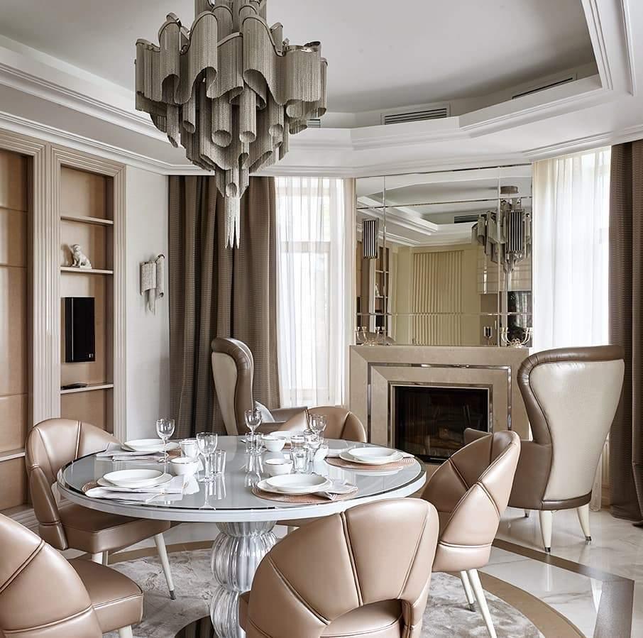A round table and fireplace sit beneath this lit silver Clérachic chandelier in a beige dining room.