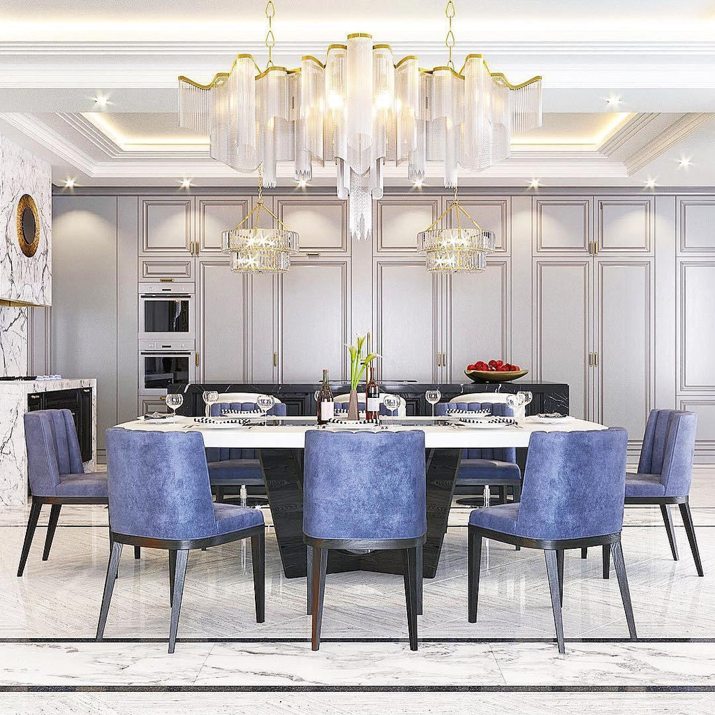 Above a long marble dining table with blue upholstered chairs, this lit Clérachic chandelier hangs with pale chain fringe and a gold-rimmed frame.