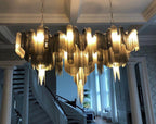 A wide gold Clérachic chandelier hangs lit above a staircase in a grand hall with coffered ceiling beams.