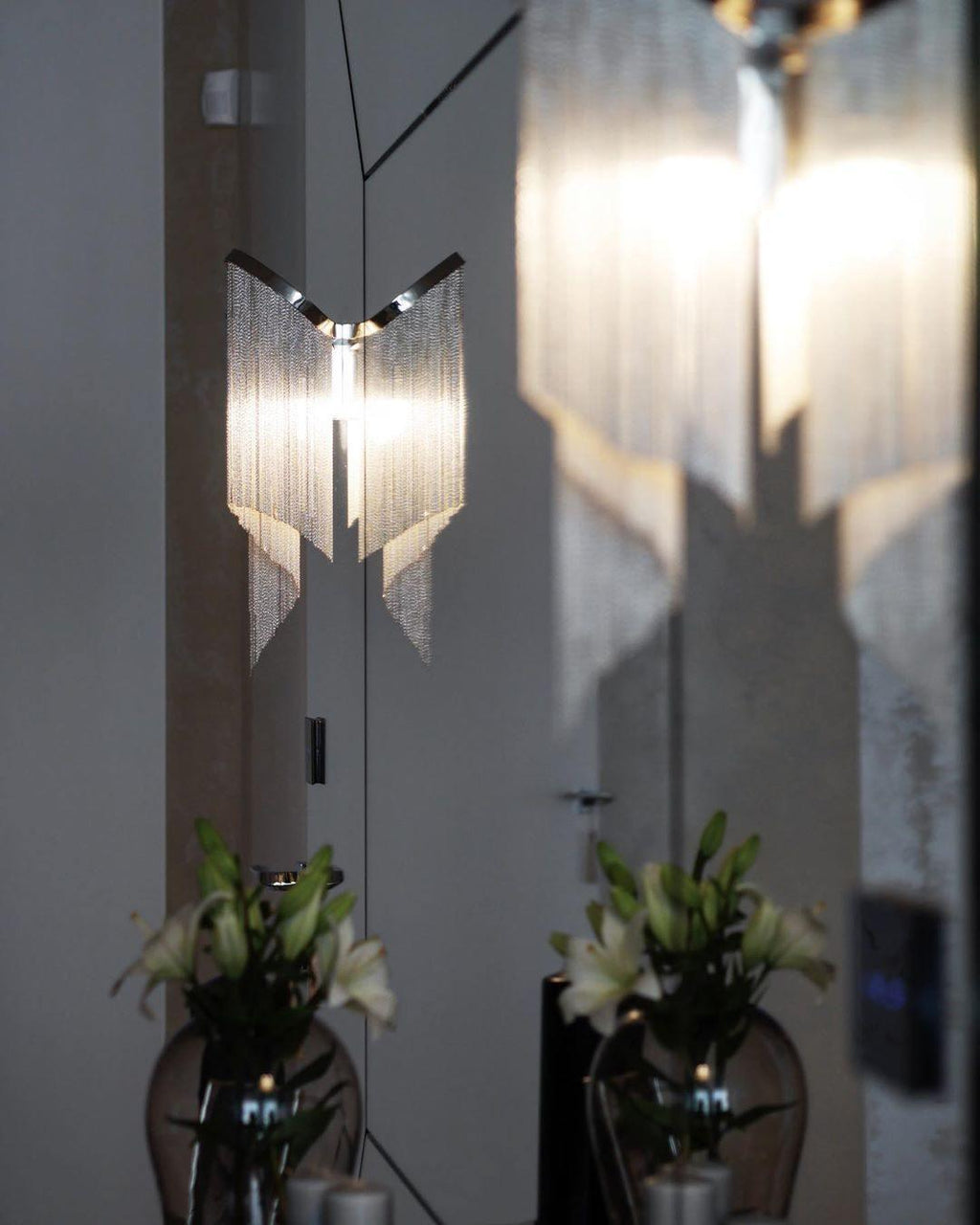 Reflected in mirrored doors, this lit silver Clérachic chandelier glows beside vases of white lilies.
