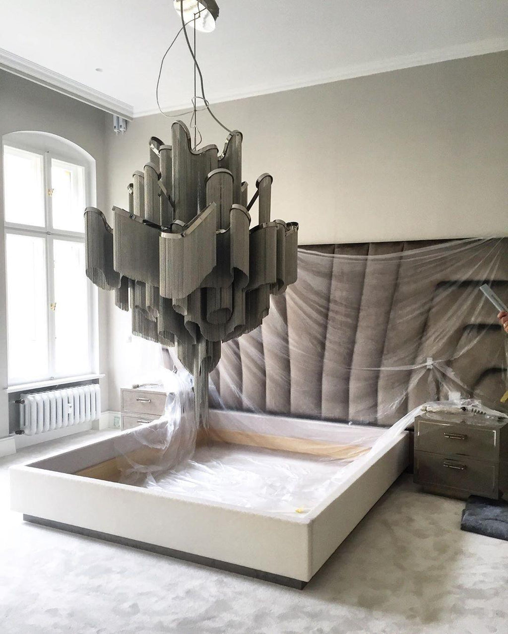A muted grey-toned Clérachic chandelier hangs over a bed with pale fabric draped across the mattress below.