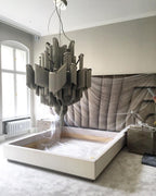 A muted grey-toned Clérachic chandelier hangs over a bed with pale fabric draped across the mattress below.