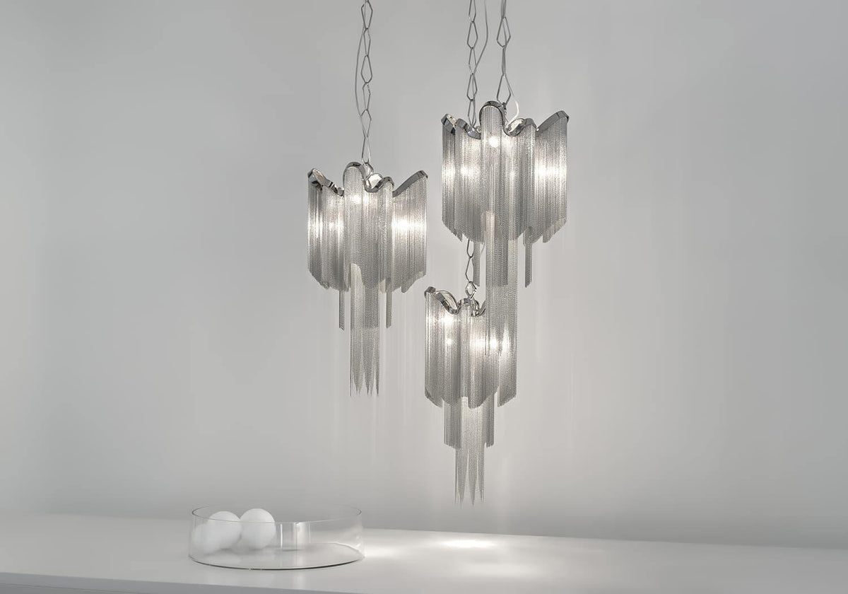 Three silver Clérachic chandeliers cluster together, lit, above small glass orbs on a plain backdrop.