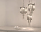 Two silver Clérachic chandeliers hang lit together against a plain backdrop, with glass orbs on the surface below.