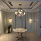 A round marble table sits beneath this lit silver Clérachic chandelier in a hallway with wall sconces either side.
