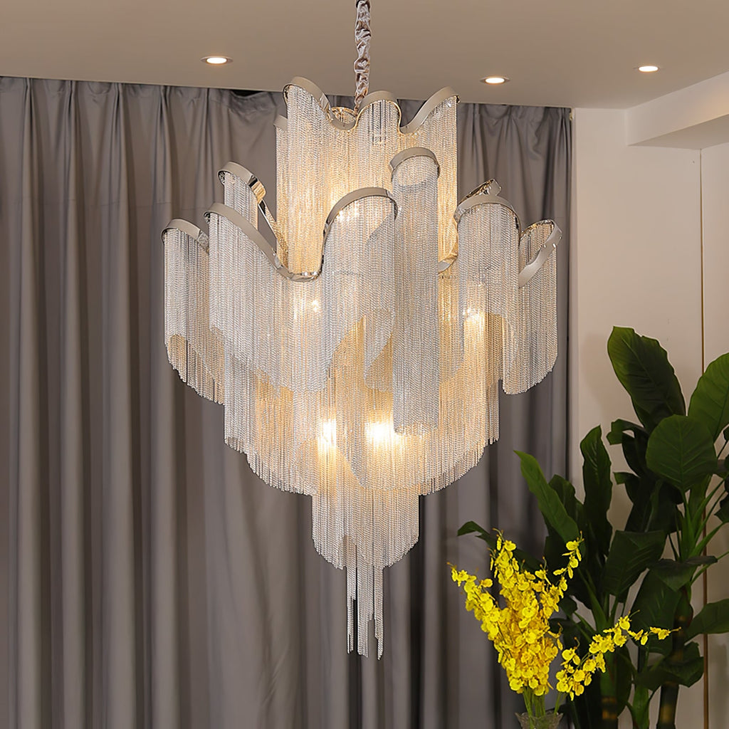 A lit silver Clérachic chain chandelier hangs above a yellow-flowered plant, framed by sheer grey curtains and cove lighting.