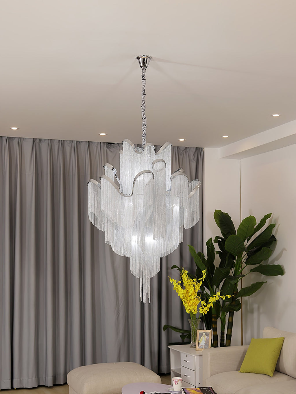 Cropped close beside a yellow-flowered plant, this lit silver Clérachic chandelier hangs against grey curtains.