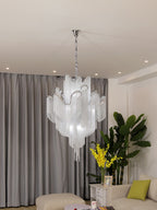 Cropped close beside a yellow-flowered plant, this lit silver Clérachic chandelier hangs against grey curtains.