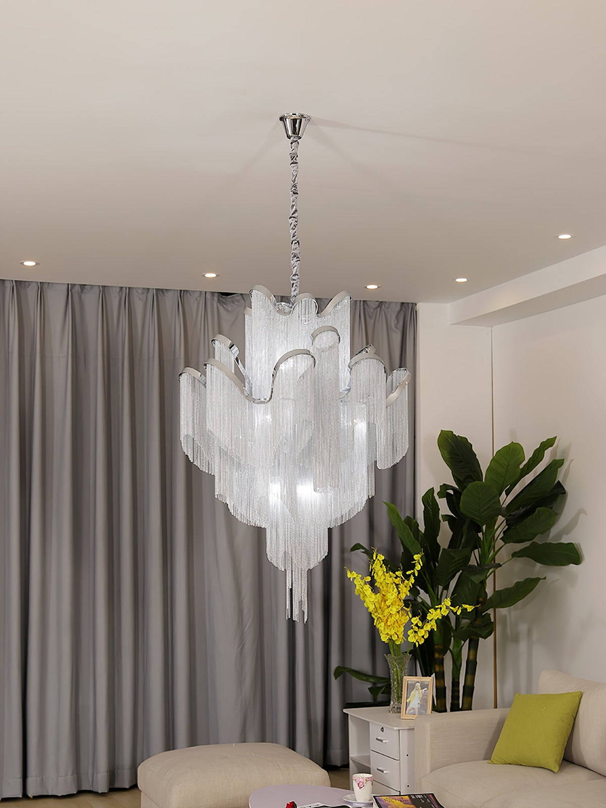 Cropped close beside a yellow-flowered plant, this lit silver Clérachic chandelier hangs against grey curtains.