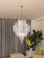 Close beside a leafy plant, this lit silver Clérachic chandelier hangs against grey curtains.