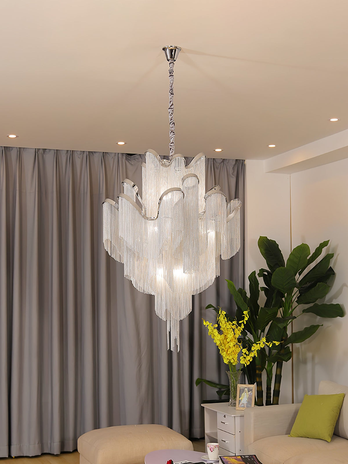 Close beside a leafy plant, this lit silver Clérachic chandelier hangs against grey curtains.