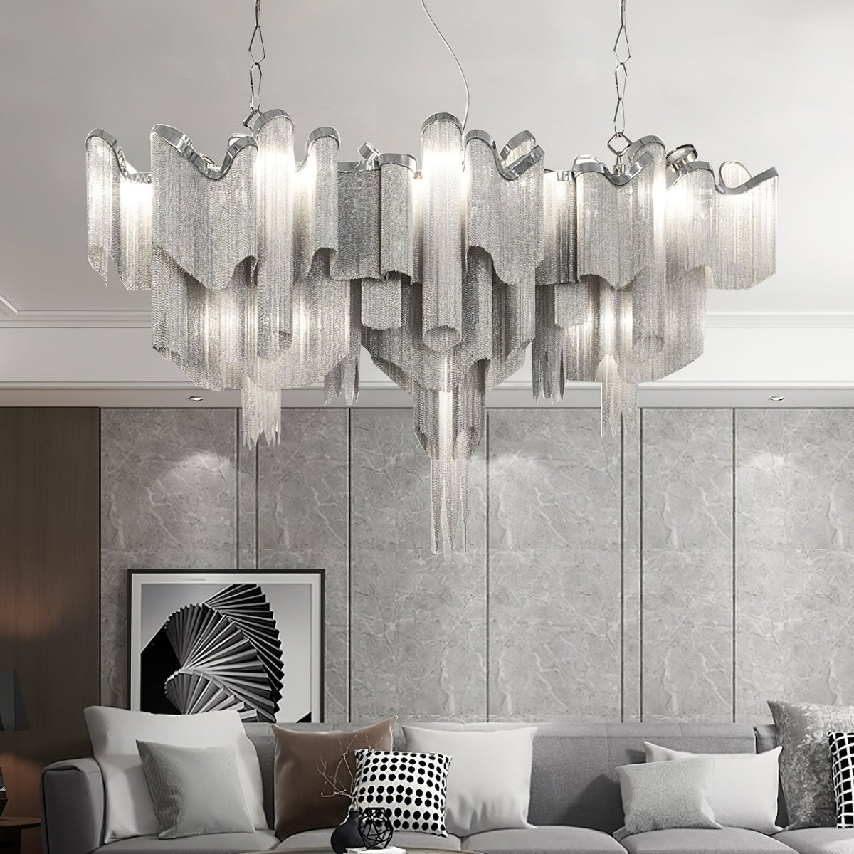 This wide silver Clérachic chandelier, lit, spans a grey panelled wall above a white sofa and patterned cushions.