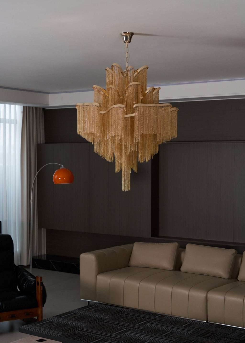 Seen from a distance, this lit gold Clérachic chandelier hangs in a lounge with tan sofas along the wall.