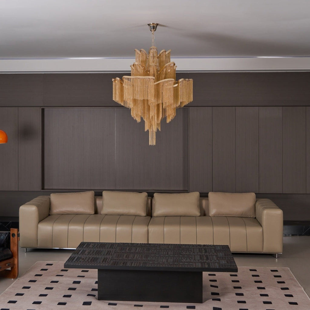 A black coffee table and long tan sofa sit beneath this lit gold Clérachic chandelier in a lounge.