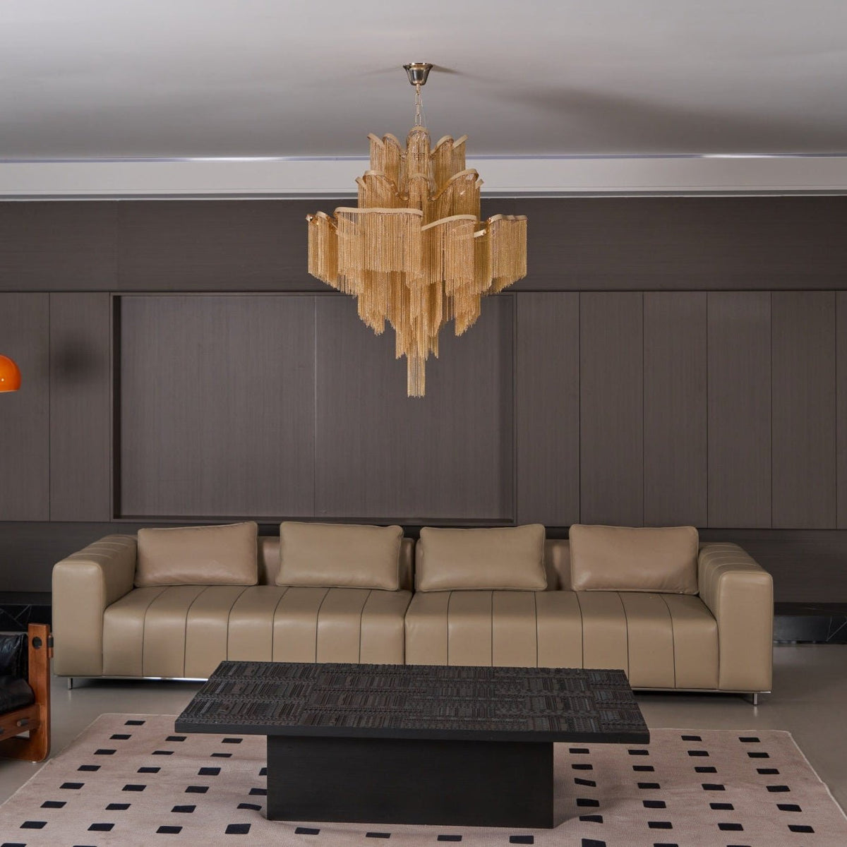 A black coffee table and long tan sofa sit beneath this lit gold Clérachic chandelier in a lounge.