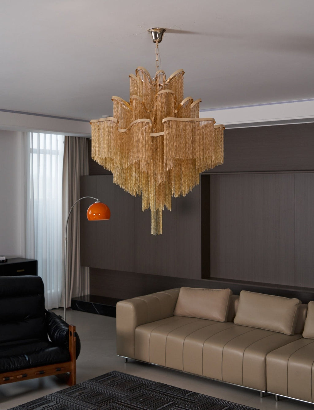 An orange arc floor lamp and black leather armchair sit near this lit gold Clérachic chandelier in a lounge.