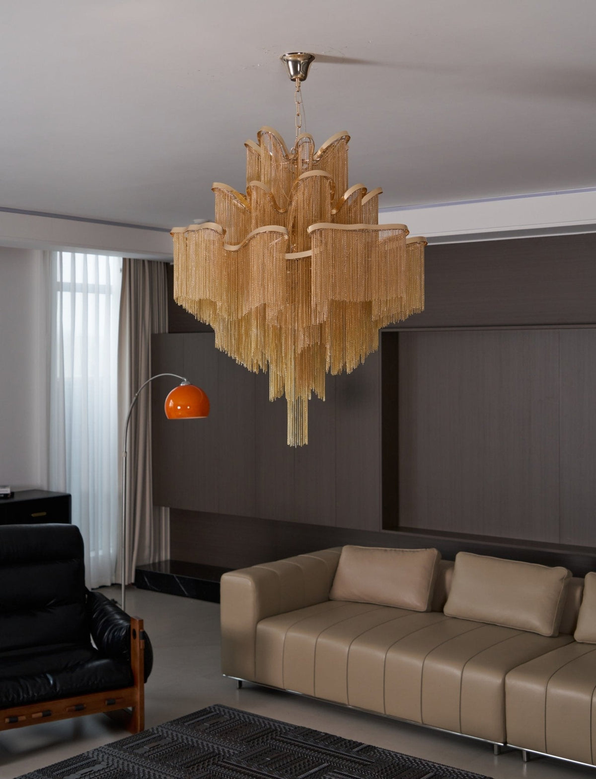 An orange arc floor lamp and black leather armchair sit near this lit gold Clérachic chandelier in a lounge.
