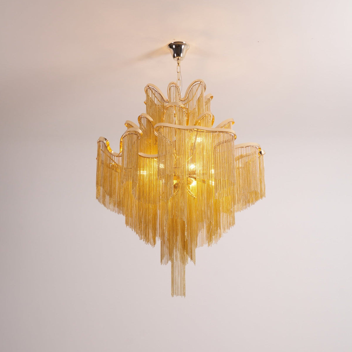 On a plain backdrop, this lit gold Clérachic chandelier shows its ceiling mount plate above the tiers.
