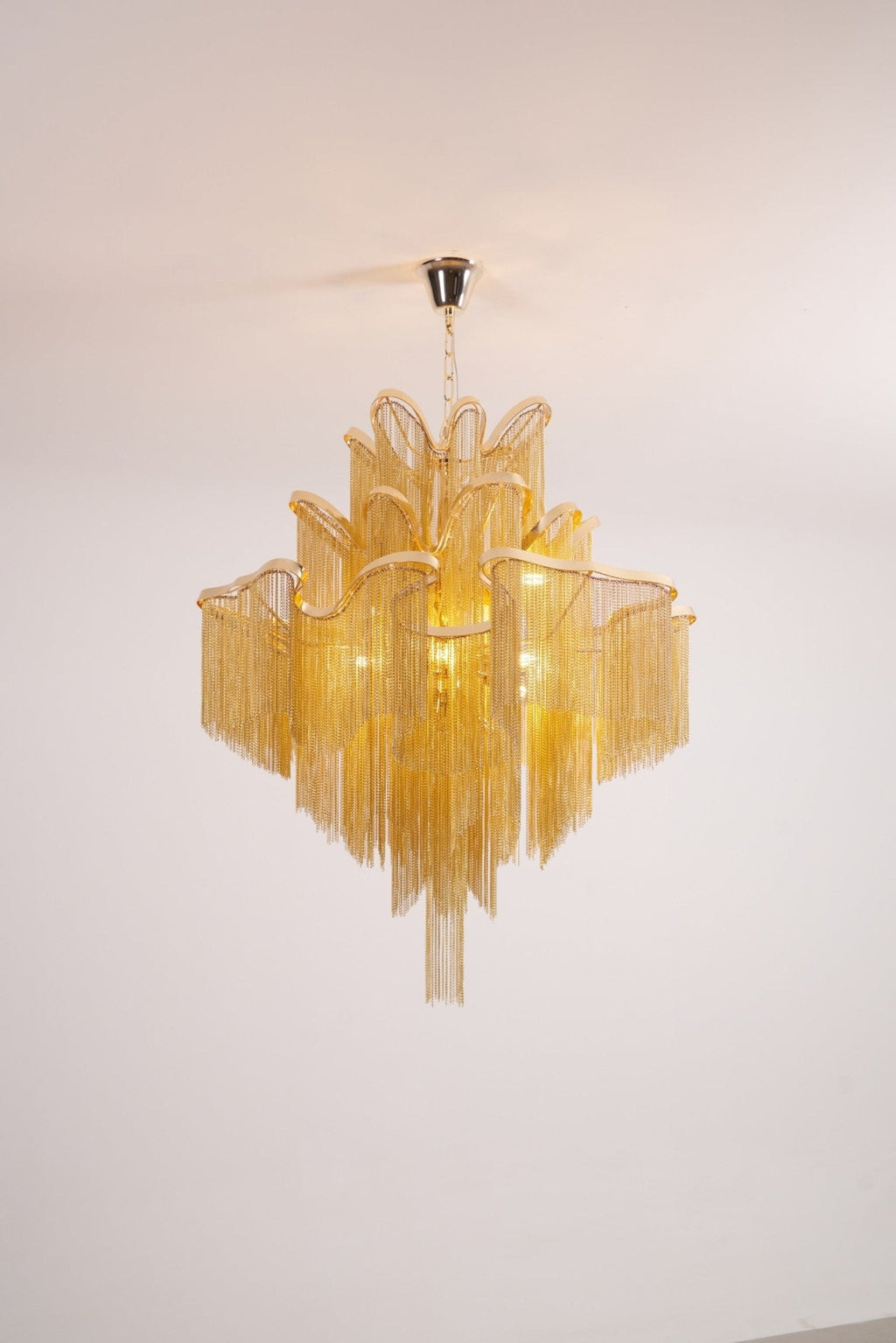 A gold Clérachic chandelier, unlit, is shown against a white backdrop with its chain hanging in tiers.
