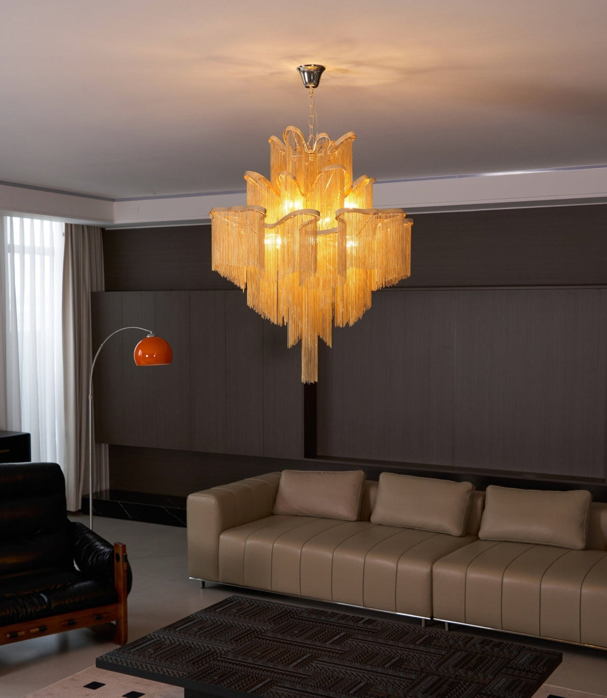 A gold Clérachic chandelier lights a lounge with tan leather sofas and an orange arc floor lamp.