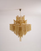 Chain fringe hangs in tiers on this lit gold Clérachic chandelier, shown against a plain backdrop.