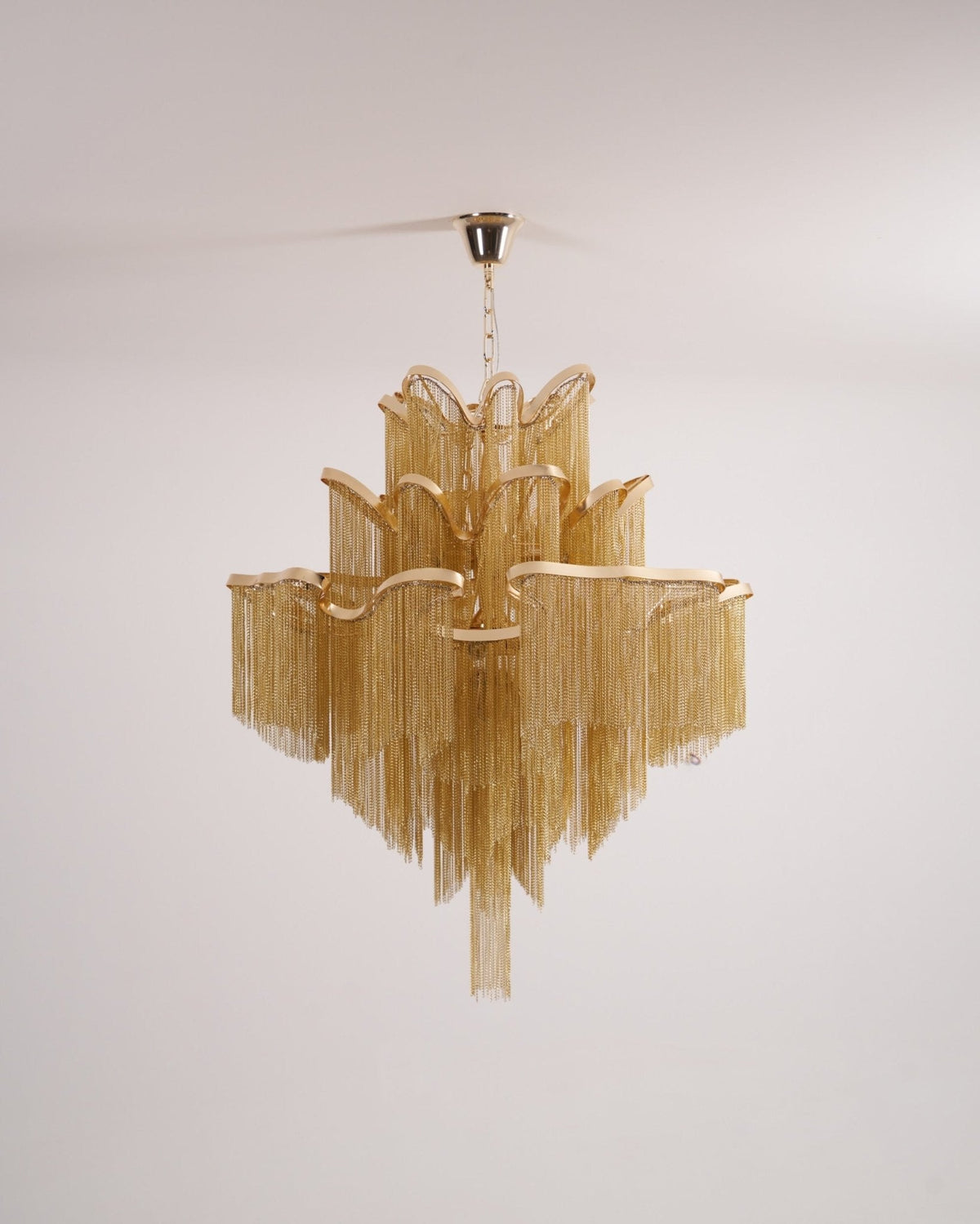 Chain fringe hangs in tiers on this lit gold Clérachic chandelier, shown against a plain backdrop.