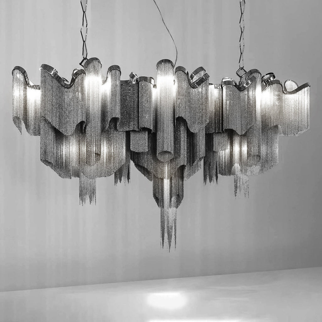Several tiers of this silver Clérachic chandelier cluster together, lit, against a plain grey backdrop.