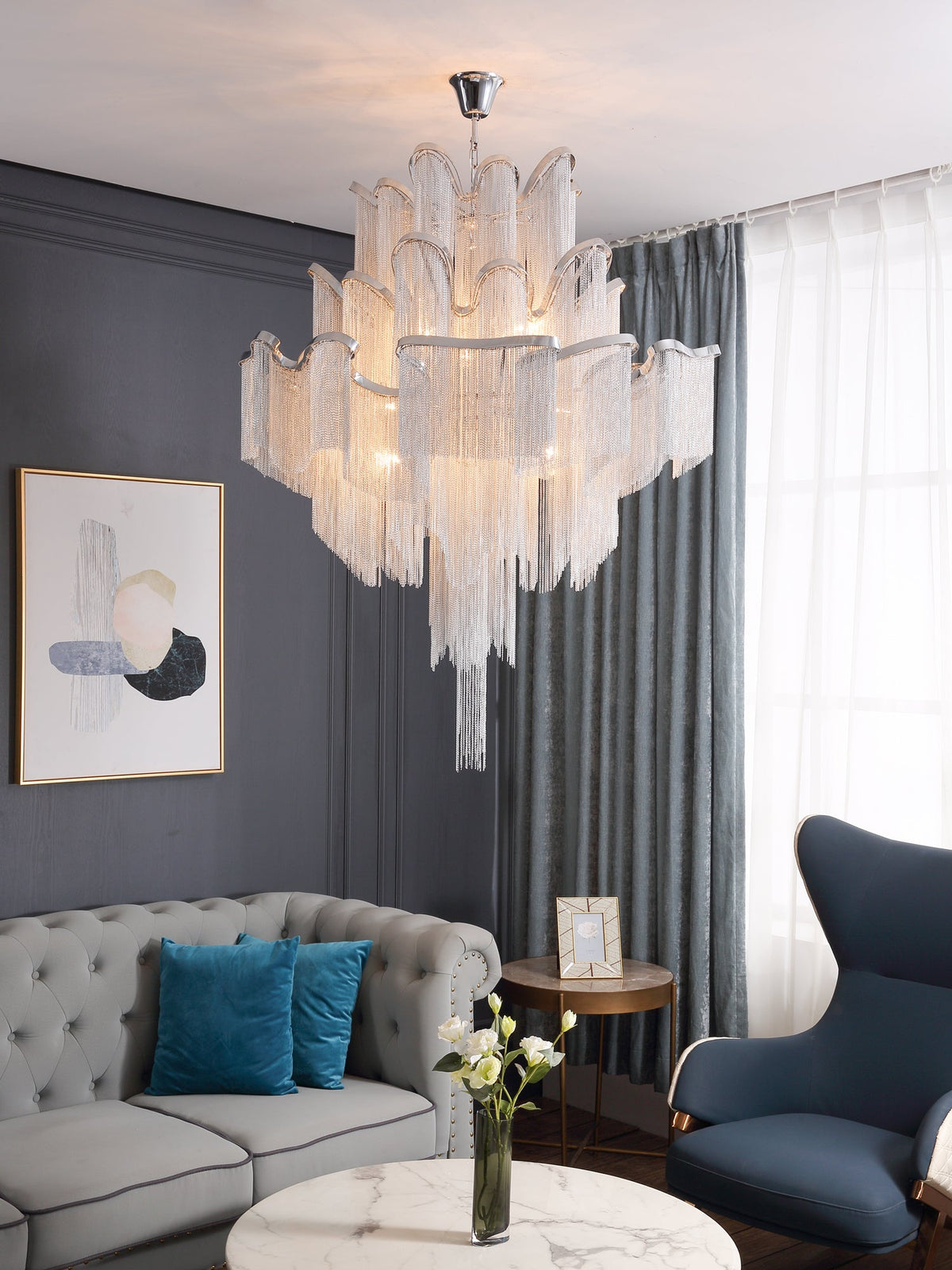 In a living room with a grey sofa and navy wingback chair, this silver Clérachic chandelier hangs lit overhead.