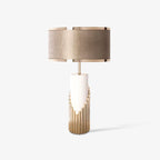 This Clérachic table lamp stands alone on a plain grey backdrop, its champagne drum shade sitting atop a stepped gold-tube base.