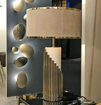 A close-up shows this Clérachic lamp's stepped gold-tube base on a black table, set against a dark mirror with sculpted spheres.