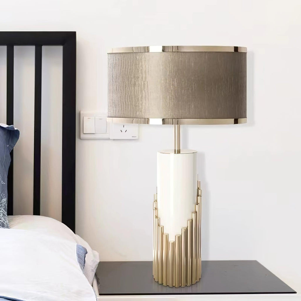 Beside a wall light switch, this Clérachic table lamp sits on a dark glass table next to a bed dressed in white linen.