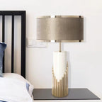 Beside a wall light switch, this Clérachic table lamp sits on a dark glass table next to a bed dressed in white linen.