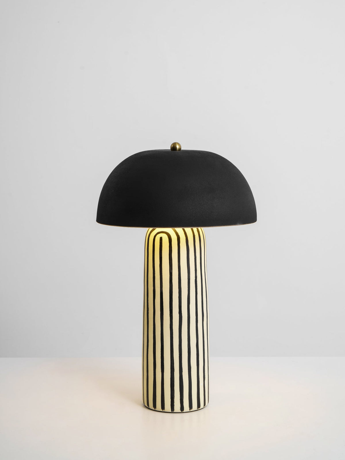 Set on a plain light surface, this Clérachic table lamp shows its striped ceramic base beneath the matte black dome.