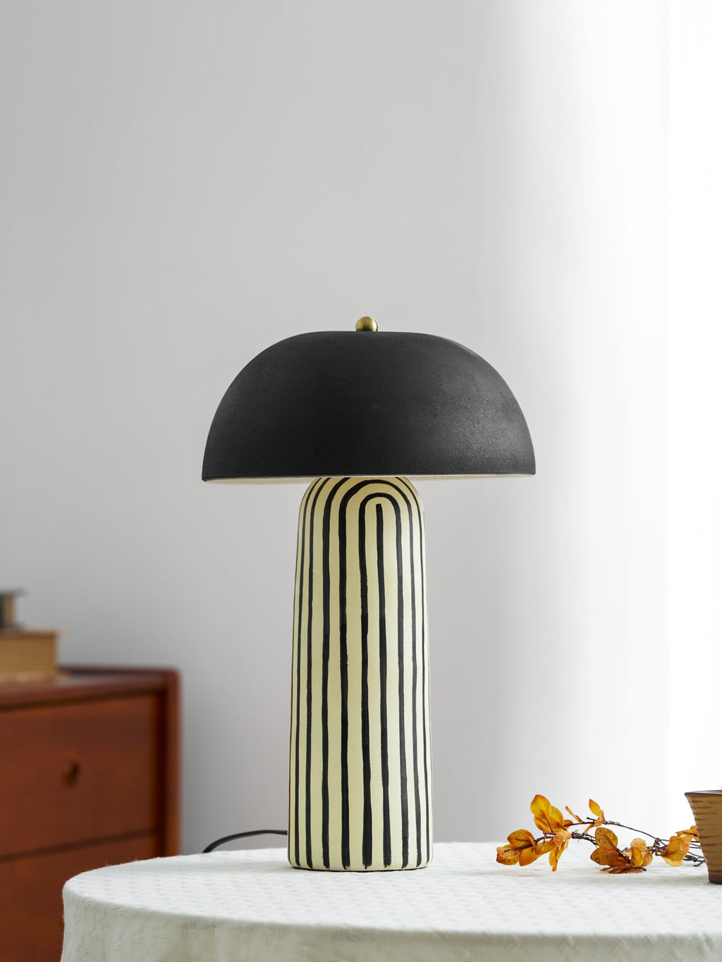 Against a plain white wall, this Clérachic table lamp stands on a round linen-covered table beside a berry branch.