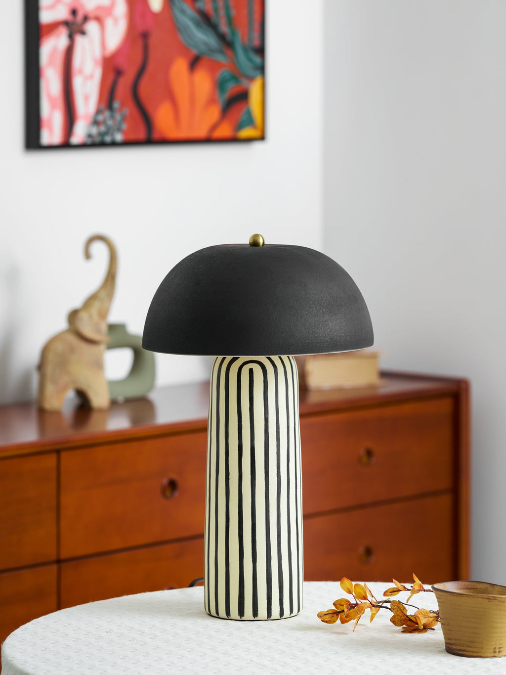 A small ceramic elephant figurine sits beside this Clérachic table lamp, a colourful painting above the dresser behind.
