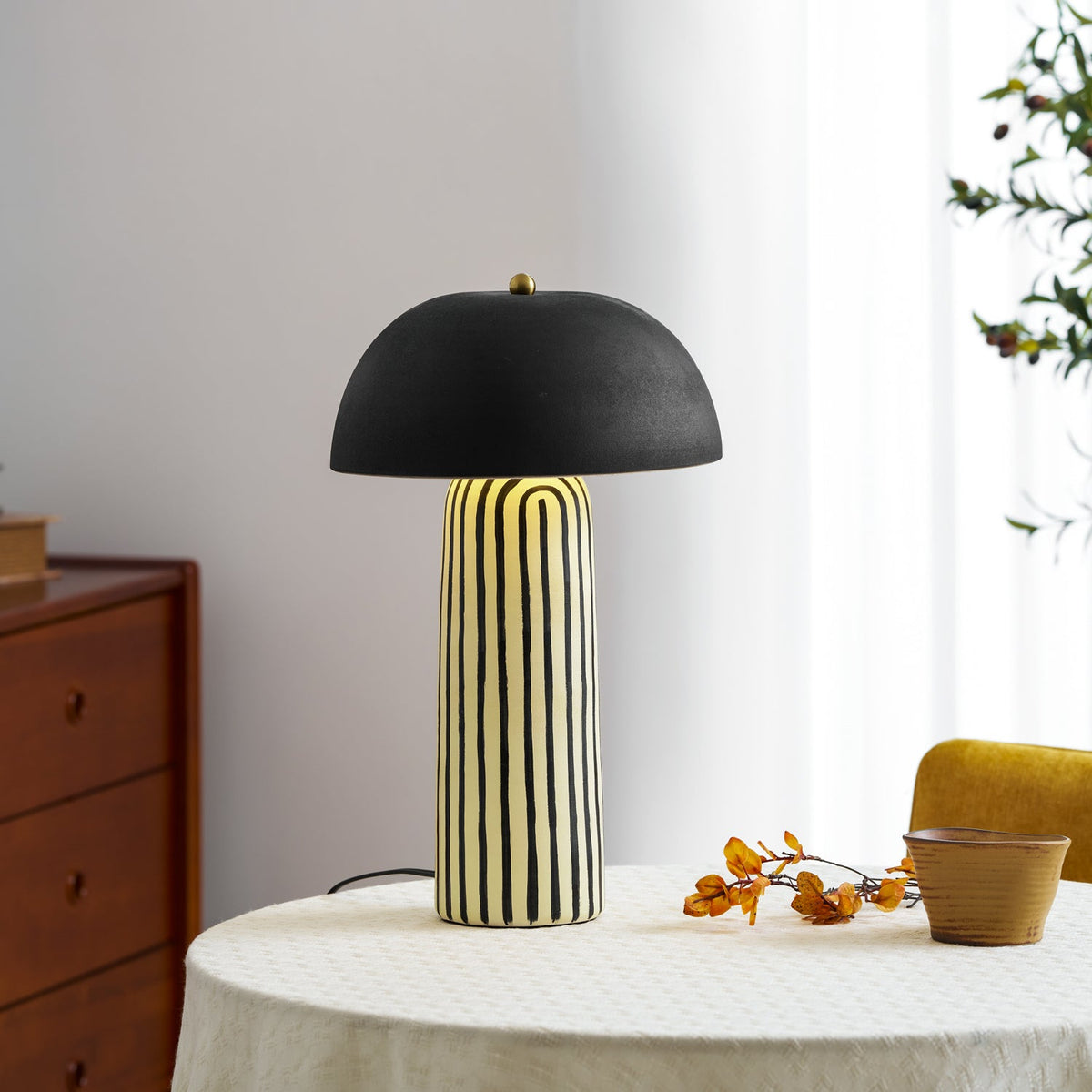 A round table dressed in cream linen holds this Clérachic table lamp, with a sheer curtain and mustard chair behind.