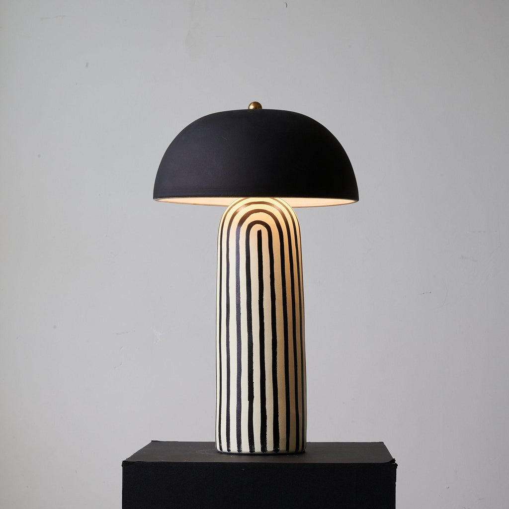 Lit on its black plinth, this Clérachic table lamp's matte black dome glows warmly above the striped ceramic base.