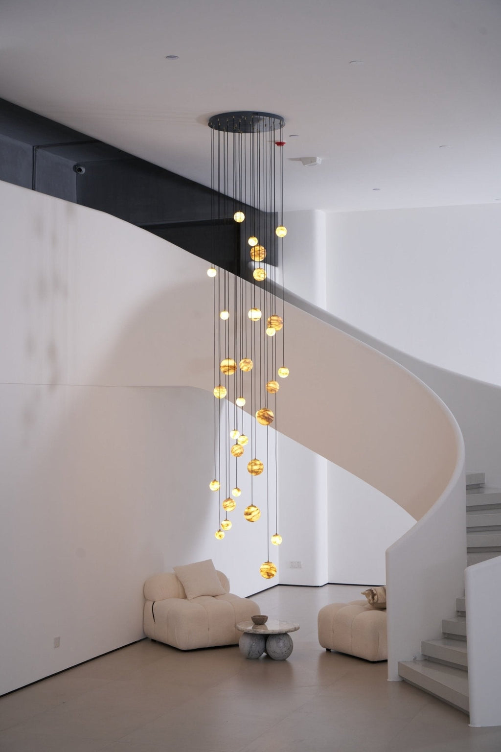 The Sultana chandelier lit in a double-height stairwell, its cascade tracing a white spiral stair.