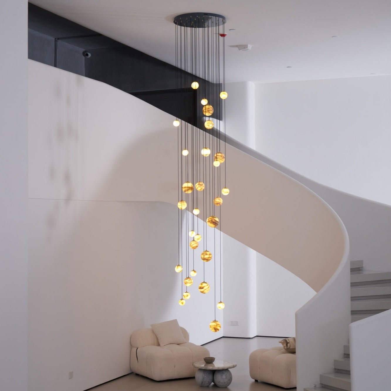 Square view of the lit chandelier filling the void beside a sculptural white spiral staircase.