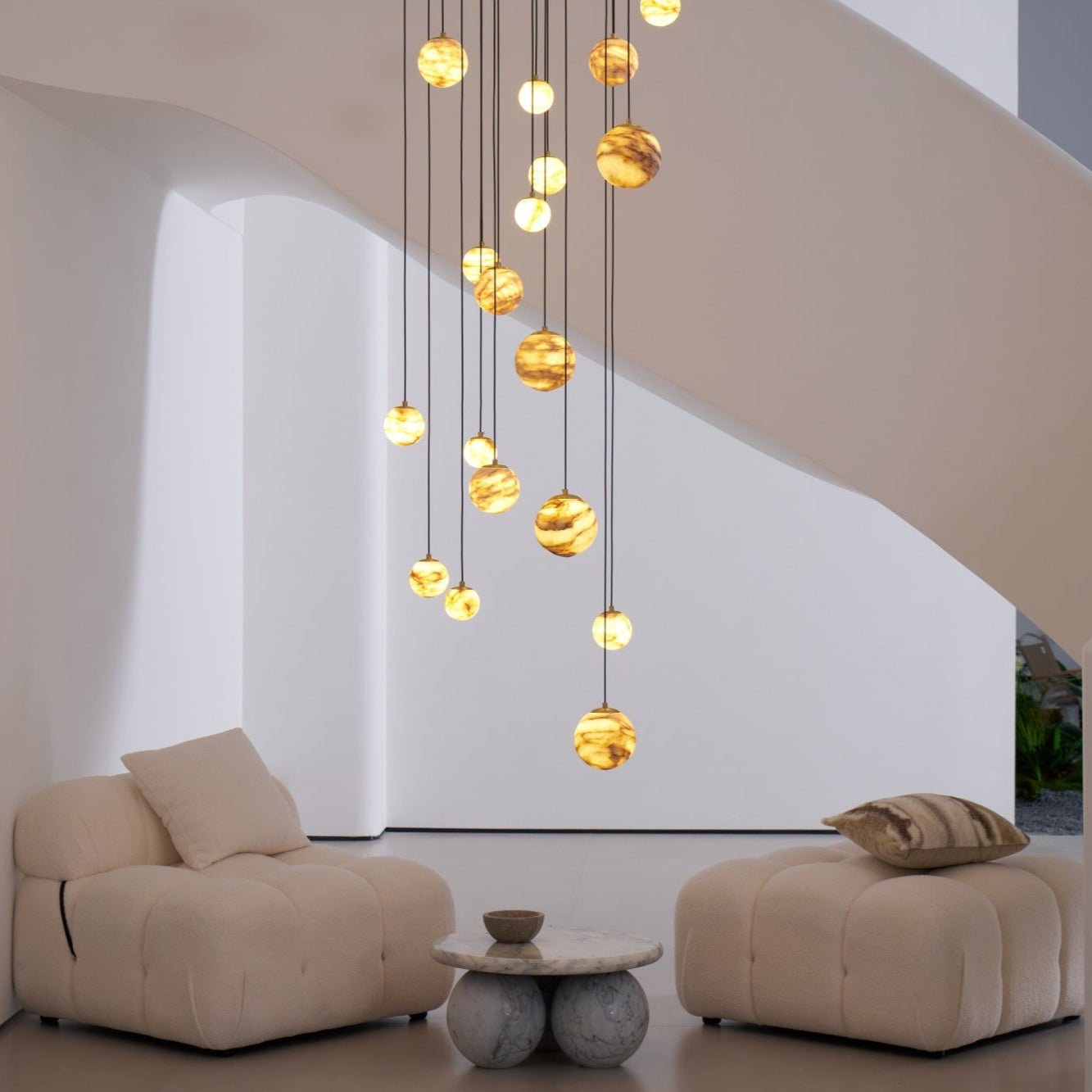 Eye-level daylight view of the lit chandelier above a marble table and pair of cream lounge chairs.