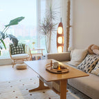 Clérachic teak floor lamp lit in a living room corner near a rattan armchair, a low coffee table, and a window with houseplants.