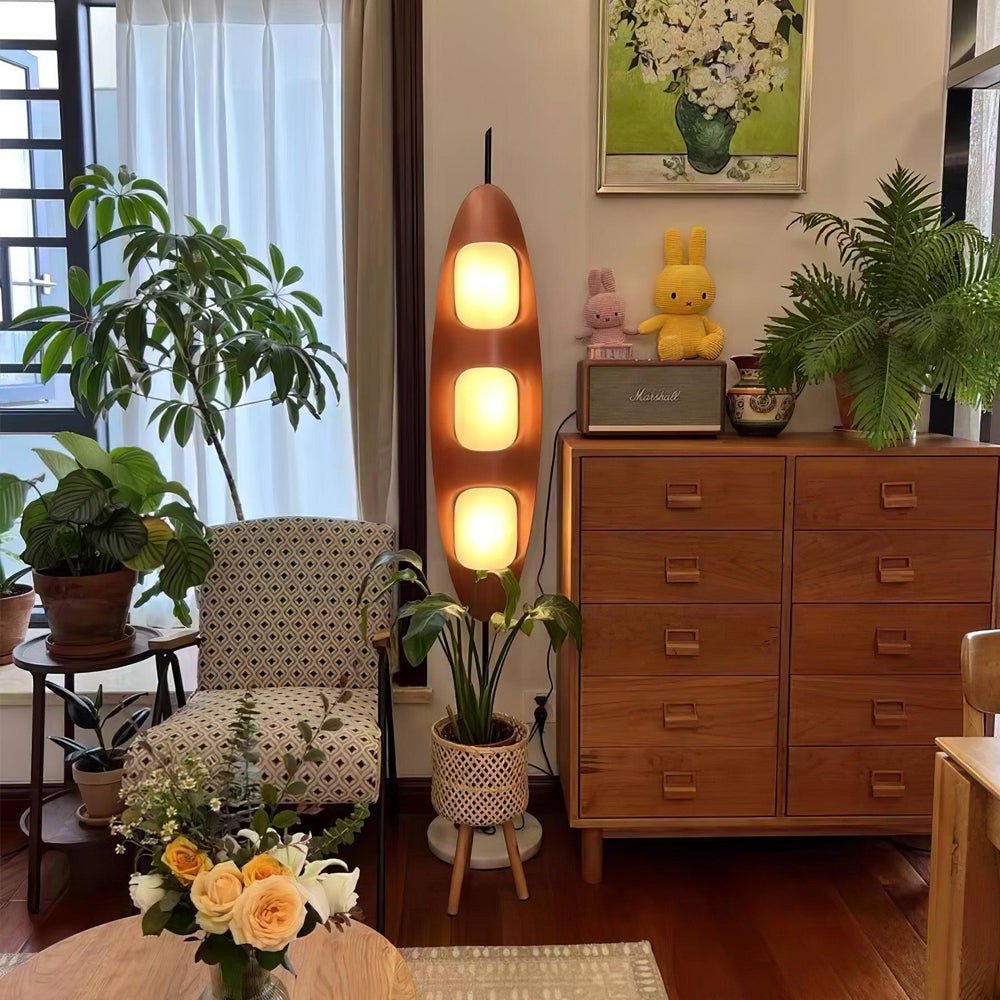 Clérachic teak floor lamp glowing beside a wicker armchair, a wooden dresser, and potted plants in a plant-filled living room corner.