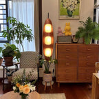 Clérachic teak floor lamp glowing beside a wicker armchair, a wooden dresser, and potted plants in a plant-filled living room corner.